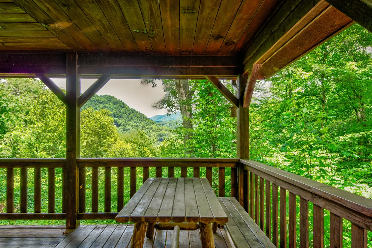 A rustic wooden picnic table sits on a wrap-around porch overlooking rolling mountains, surrounded by lush greenery. The setting is perfect for outdoor dining, morning coffee, or simply savoring the peace of nature. The sturdy wooden railing and wide porch emphasize the indoor/outdoor connection.