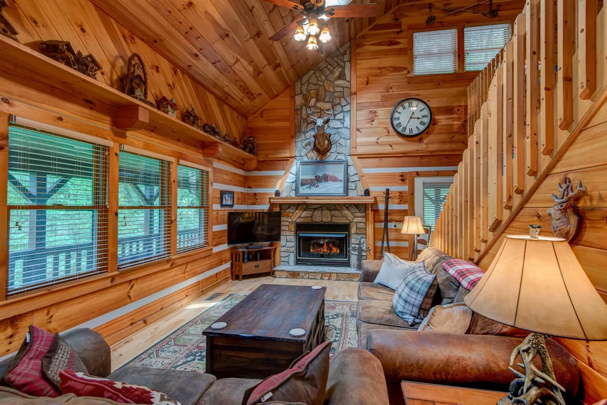 A spacious living room featuring a grand stone fireplace, rustic wooden beams, large windows that flood the space with natural light, and comfortable, inviting sofas and chairs. The decor is cozy and mountain-inspired, blending natural materials to create a welcoming gathering area.