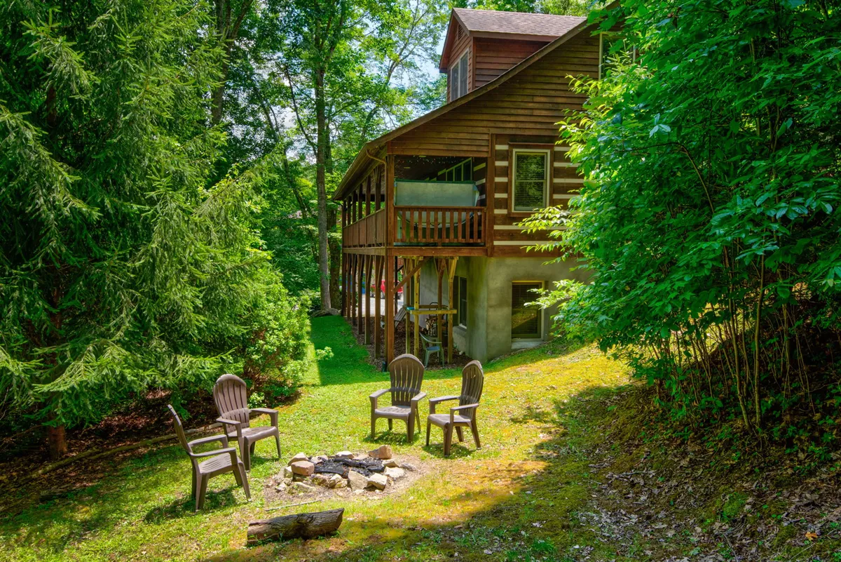A cozy fire pit ringed by wooden chairs sits just off the deck, surrounded by tall trees and the soft glow of evening light. The deck features easy access to the backyard, inviting guests to gather, share stories, toast marshmallows, and enjoy the tranquil embrace of nature.