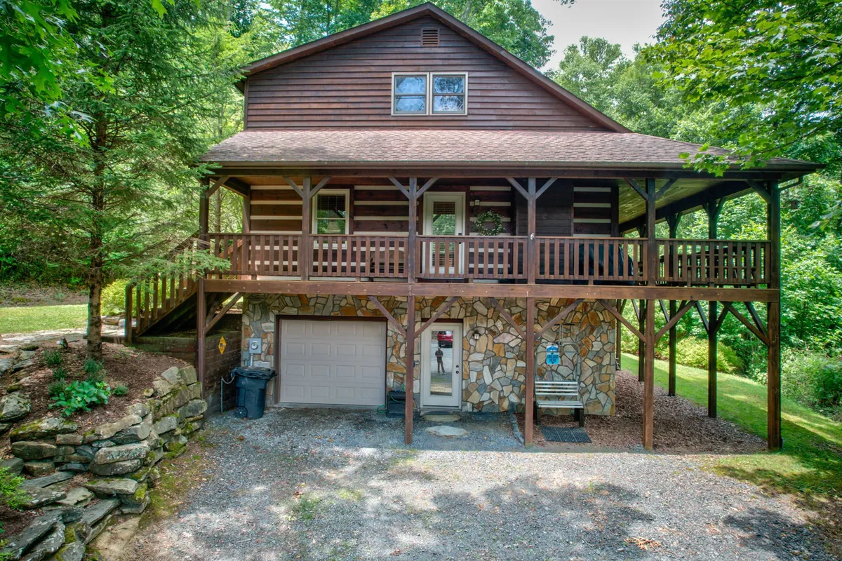 A broad wooden deck and stone facade welcome guests at the entrance, surrounded by leafy trees. The spacious porch area suggests ample room for group gatherings and outdoor activities, with a warm, inviting atmosphere.