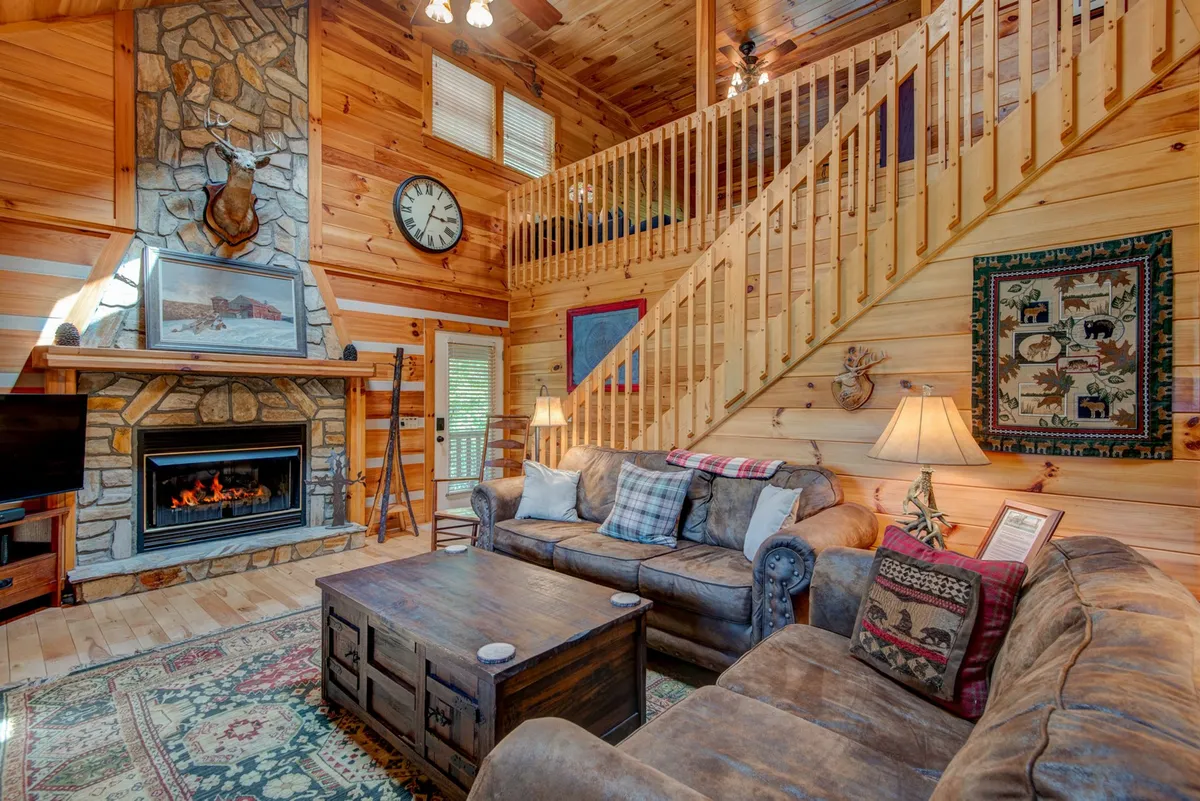 An inviting downstairs living area with a prominent stone fireplace, cozy sofas clustered for conversation or TV viewing, a wooden staircase, and warm lighting. The space exudes comfort and is suited for both quiet reading and lively family gatherings.