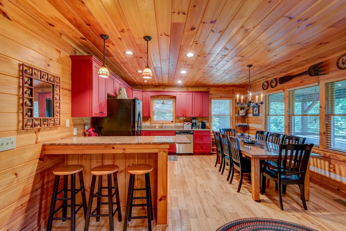An open-concept kitchen and dining area with red cabinetry, a kitchen island with granite countertops, and a spacious dining table set for a group. The layout encourages socializing and effortless flow between cooking and dining.