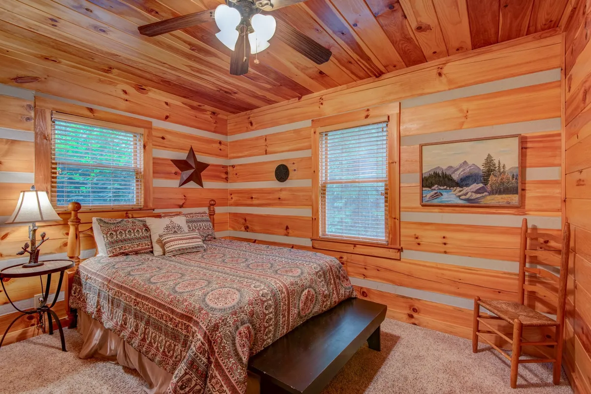 A welcoming bedroom with patterned bedding, wooden walls, and ample natural light streaming through windows with blinds. A decorative star and a serene painting add personalized touches, conveying comfort and tranquility.