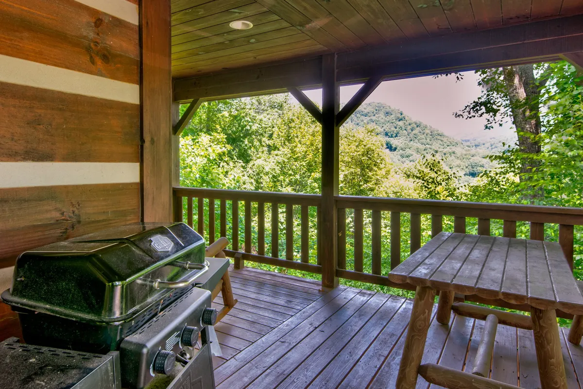 A grill and wooden table are set on a scenic deck with a panoramic mountain backdrop. The scene suggests a welcoming space for sunset barbecues, morning coffee, or casual meals, seamlessly blending cooking, dining, and those epic mountain views.
