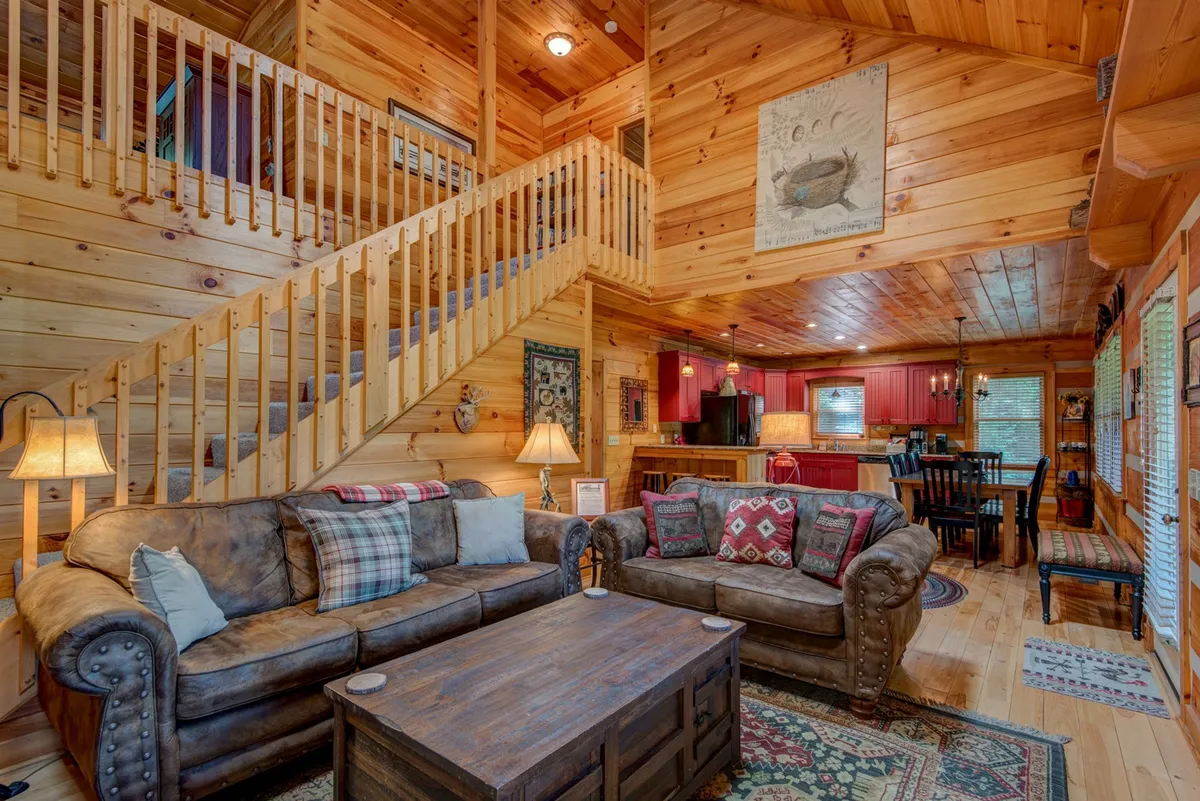 A wide-angle view of the main living area, including comfortable sofas, a rustic coffee table, a wooden staircase, and a visible kitchen area in the background. The open-plan design invites connection between guests whether lounging or preparing meals.