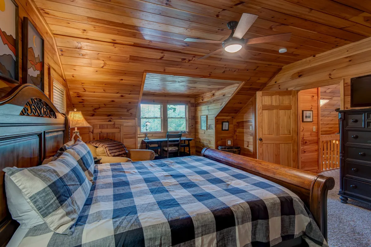 A spacious suite featuring a large bed with plaid bedding, warm wooden paneling, a decorative lamp, and a small dining area. The setup suggests a serene, self-contained space perfect for relaxation and privacy, likely representing the downstairs king suite.