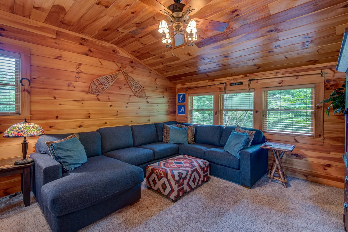 This image features a spacious sectional sofa, a colorful ottoman, a lamp, and wood-paneled walls with windows letting in natural light. The scene is inviting and clearly designed for relaxation, socializing, reading, or playing games.